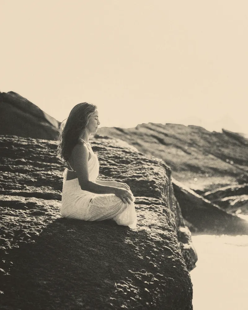Helena Alves meditando sentada sobre uma pedra junto ao mar, em preto e branco.
